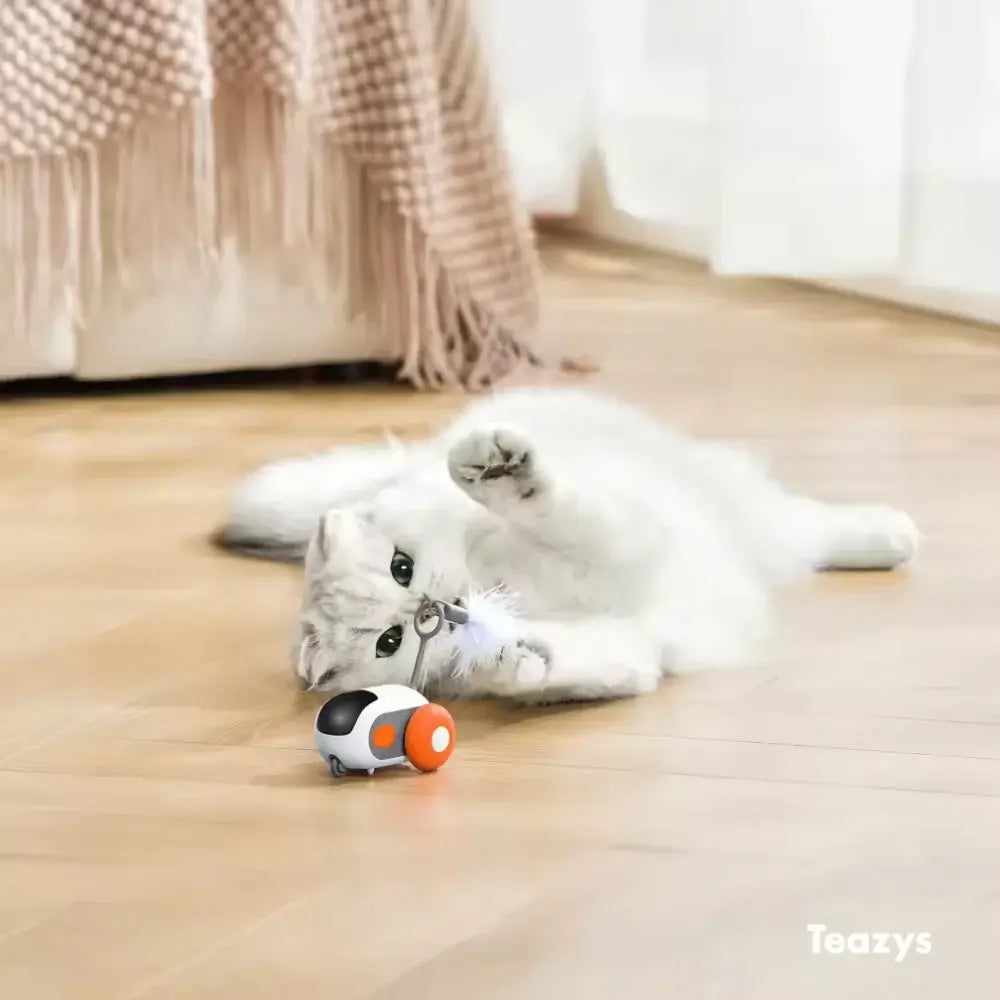 A fluffy white cat with a TURBO TAIL 2.0 rests on a wooden floor, batting at a toy spaceship. A plaid blanket is draped over the nearby sofa.