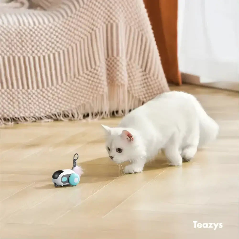 A cat with a Turbo Tail 2.0 in white curiously approaches a small toy on a wooden floor near a textured blanket.